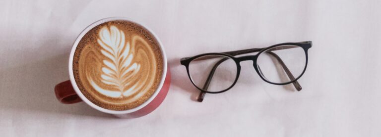 A black, round glasses frame lying next to a cup of coffee. The surface of the coffee shows latte art in the shape of a leaf.