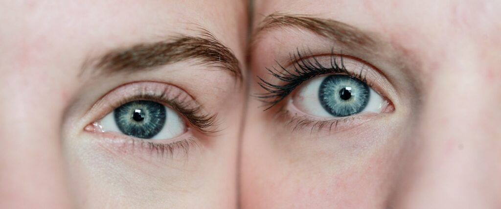 Close-up of two people with blue eyes, with the sides of their faces pressed right against each other. The left one's eye is a dark blue, the right one a brighter, light blue.