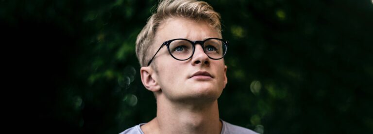 A blond man with blue eyes who is wearing black round glasses looks up at the sky.