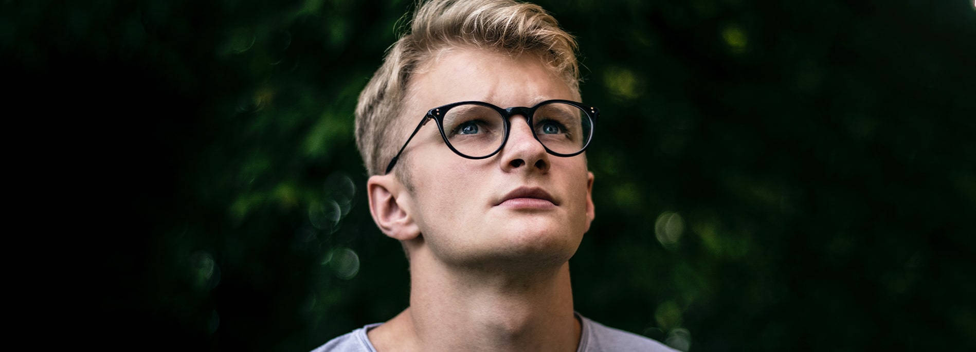 A blond man with blue eyes who is wearing black round glasses looks up at the sky.