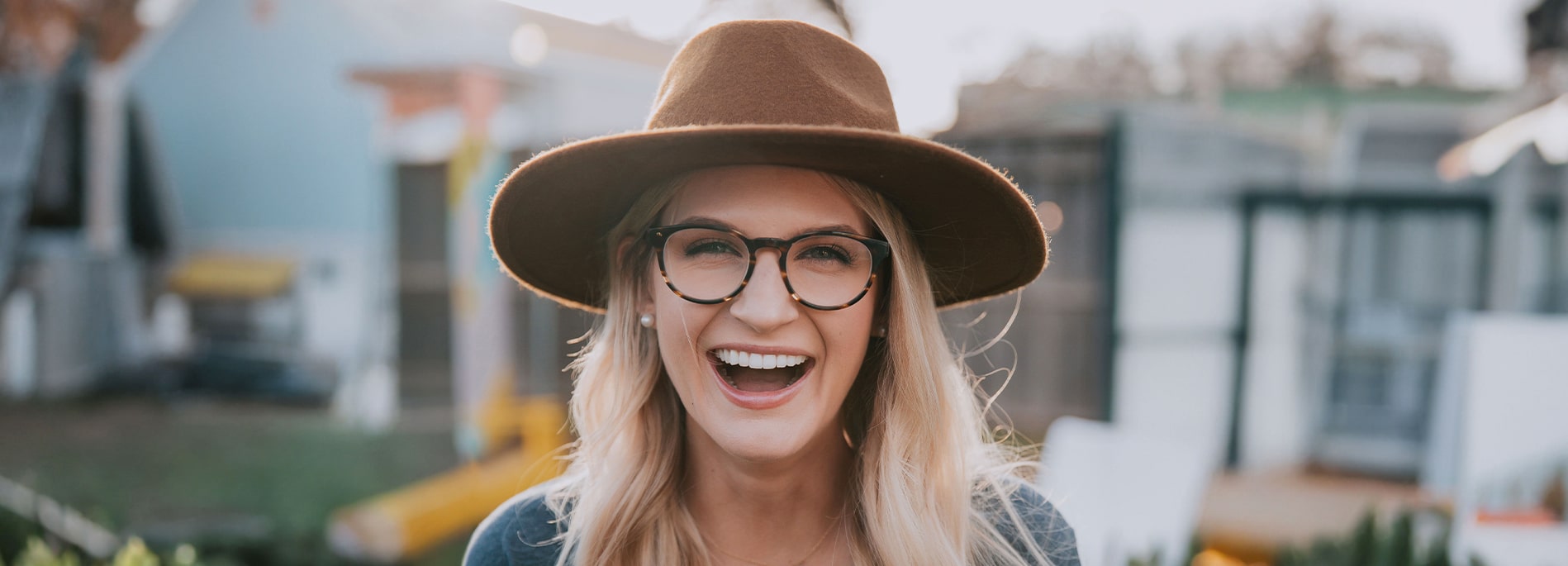A laughing blonde woman wearing a hat and round tortoiseshell glasses