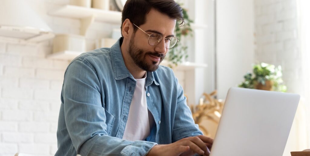 Male wearing glasses sitting at a table working on this laptop