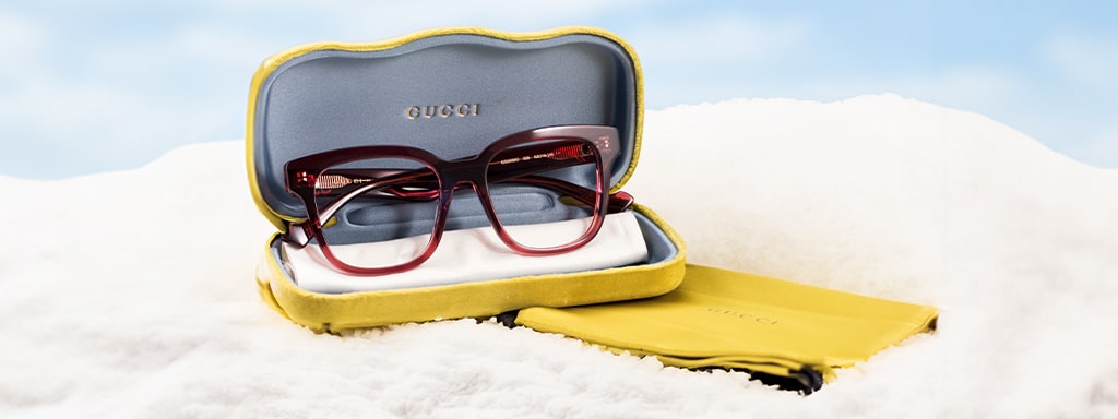 A square, red pair of Gucci glasses with yellow branded case and pouch lying on a snowy background