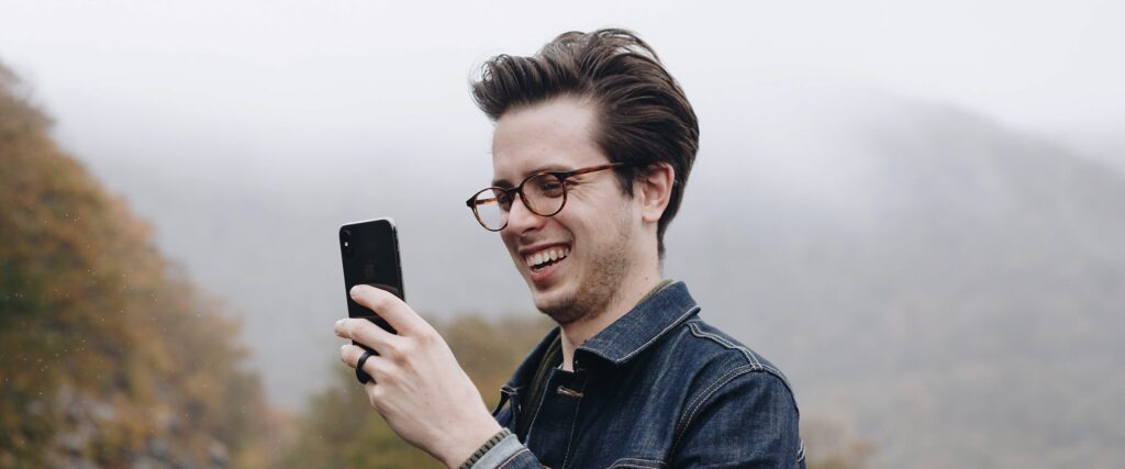 Man outdoors wearing round glasses posing for a selfie laughing