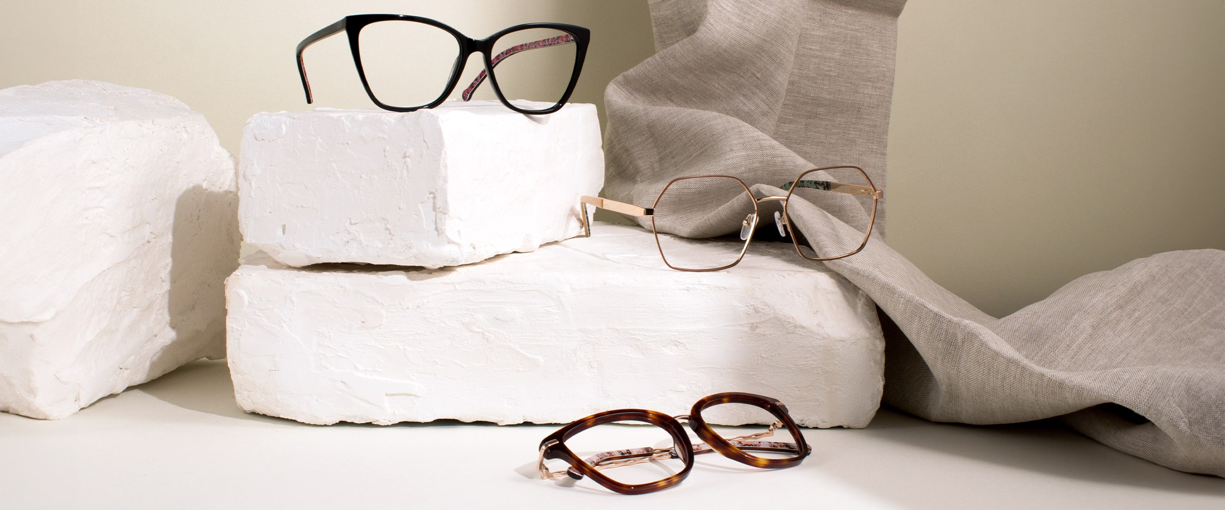 Three pairs of Scout Made in Italy frames, one tortoiseshell, one gold metal and one black positioned on different levels amongst stone blocks and fabric