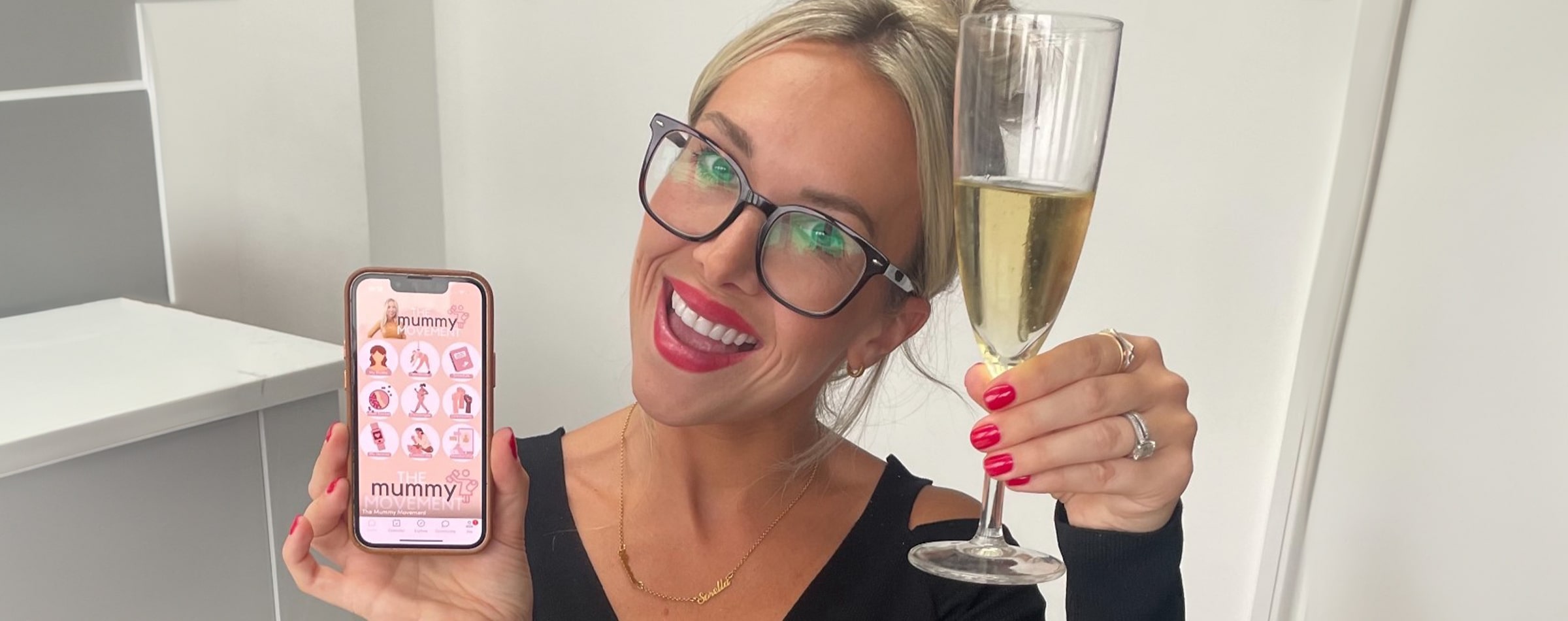 Happy woman wearing a pair of rectangular frames holding a glass of bubbles and showing her mobile phone screen of her app 'The mummy Movement'