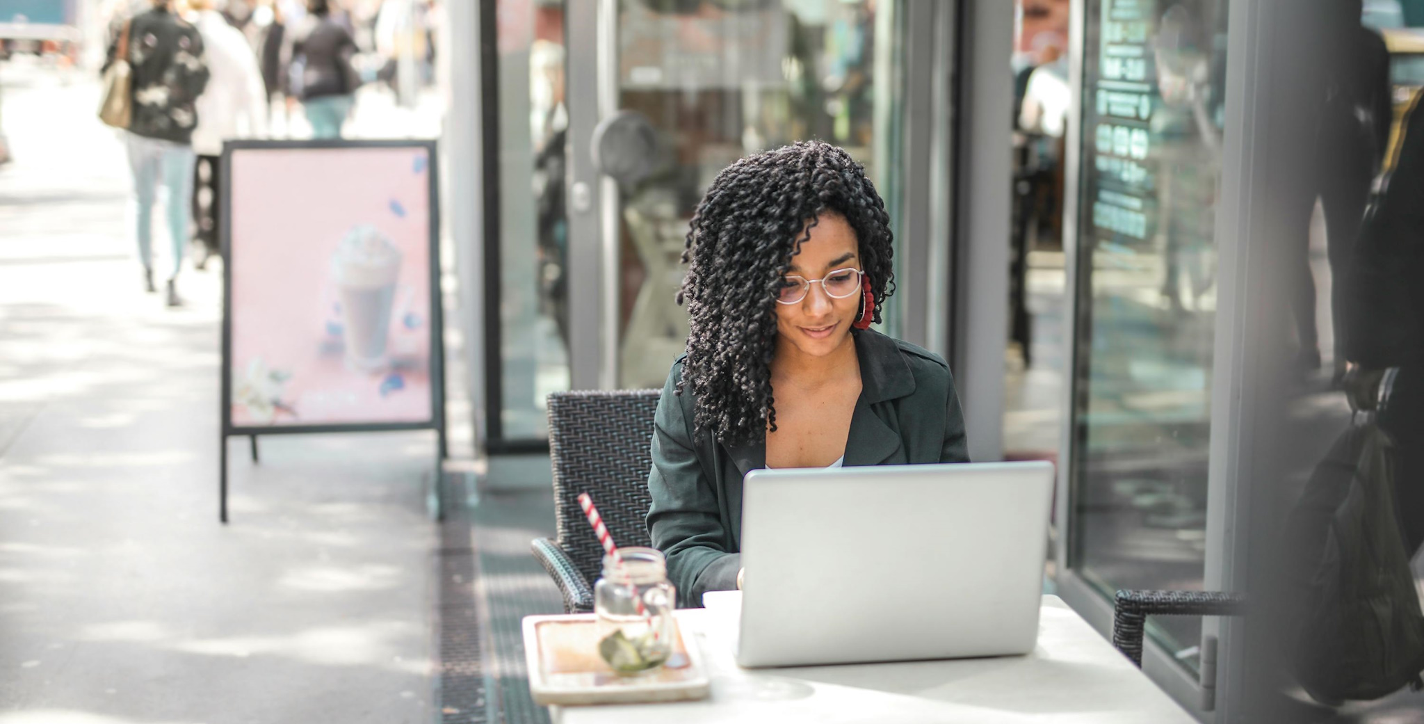 A young black woman wearing glasses sits outside a cafe working on her laptop. Her hair is in long curly ringlets and she's smiling.