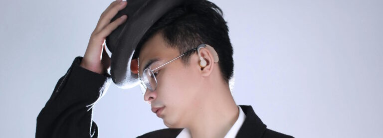 An Asian man who wears glasses and a hearing aid putting on a hat