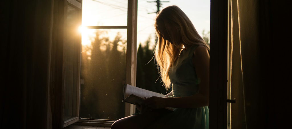 A blonde woman reading a book sitting at an open window while the sun is setting outside