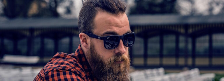 Man with a thick beard and a fauxhawk wearing Ray-Ban sunglasses while sitting outside