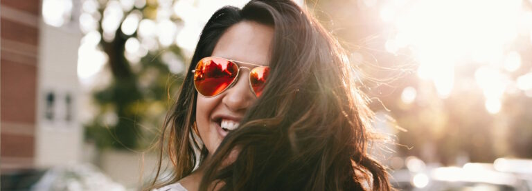 A smiling woman wearing red mirrored aviator sunglasses in the sun while her hair whirls around her head