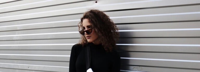 A woman with brown curly hair and wearing slim cat-eye sunglasses standing in front of a garage door