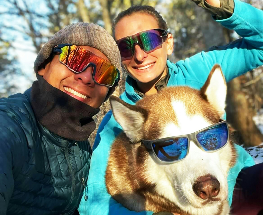 A similing man and woman pose for the camera outdoors while hugging their dog, a husky. Both the couple and the dog are wearing sporty sunglasses.
